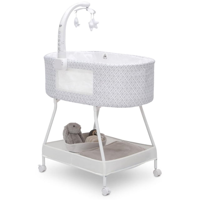 Delta Children Sweet Slumber Bassinet, Garden Path, oval bassinet with white and grey patterned fabric and breathable mesh window, featuring an electronic mobile with hanging plush toys, supported by white metal legs with caster wheels and a lower storage shelf holding a grey stuffed bunny and baby blanket.