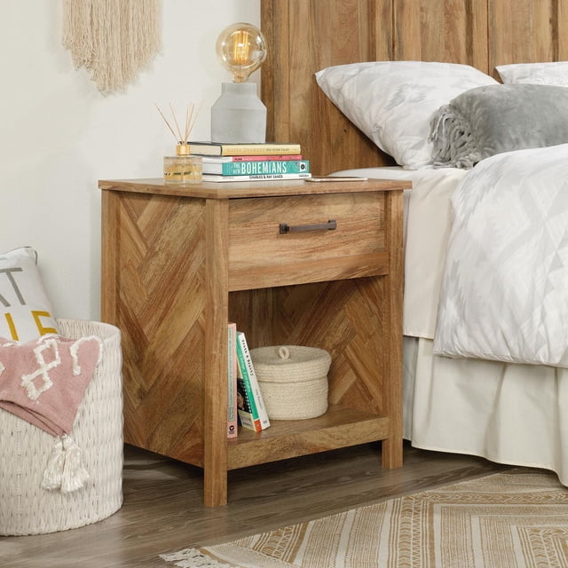 Sauder® Cannery Bridge Night Stand in natural wood finish with a drawer featuring metal runners and safety stops, an open shelf holding books and a woven basket, styled with a concrete base lamp, reed diffuser, and books beside a bed with white geometric bedding on a wood floor with a patterned rug.