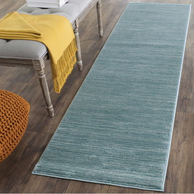 Safavieh Vision Adrasteia Overdyed Solid Area Rug in muted teal blue with subtle horizontal texture, laid on medium brown wood flooring beside a gray cushioned bench with a mustard yellow fringed throw and an orange knitted pouf, ideal as an accent rug for contemporary indoor spaces or hallways.