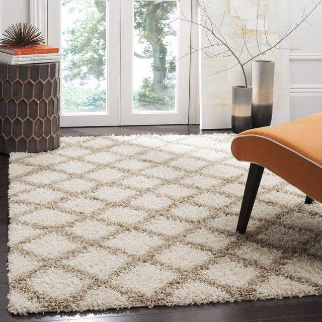 Safavieh Layla Geometric Plush Shag Area Rug featuring a cream and beige diamond pattern with dense, soft synthetic fibers, displayed on dark hardwood flooring in a contemporary room with an orange upholstered chair, a hexagonal side table with books and decor, and two gradient ceramic vases holding bare branches near glass doors, suitable for high-traffic home or office areas.