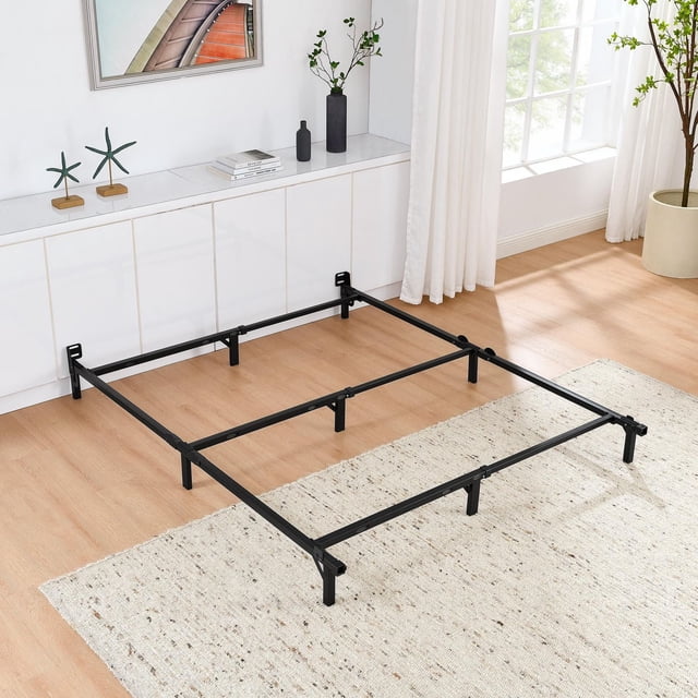 Mainstays 7" Twin/Full/Queen Size Adjustable Metal Bed Frame in black with welded legs, integrated middle support beam, built-in headboard brackets, and capped feet to protect floors. Positioned on a light wood floor with a beige rug in a bright room featuring white walls, a large window with white curtains, a white cabinet with decor, and a potted plant. Adjustable size suits guest or master bedrooms; box spring and mattress required, not included.