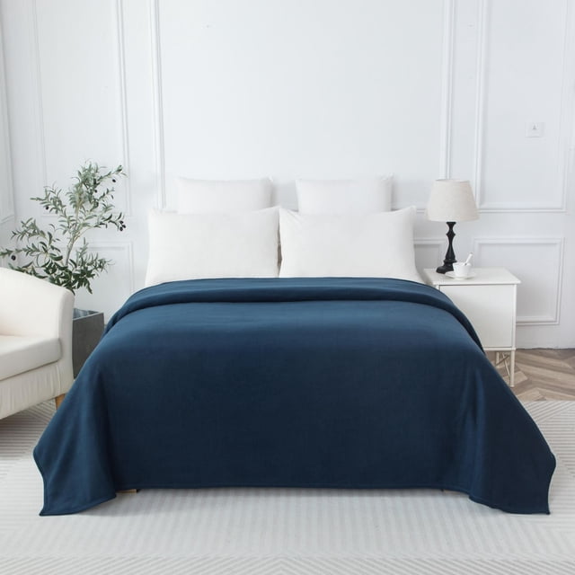 Mainstays Microfleece Blanket Blue T in deep blue polyester fabric spread over a bed with white pillows in a bright, minimalist bedroom featuring a white armchair, potted plant, and nightstand with lamp, highlighting its soft, cozy texture and warmth ideal for bedding use.
