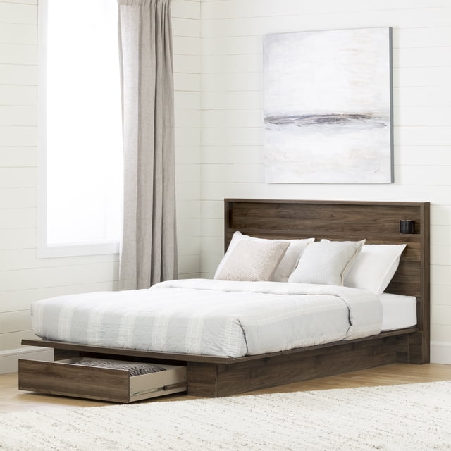 South Shore Tao Full/Queen Platform Bed and Headboard Set in natural walnut with a minimalist design, featuring a low-profile frame and a rectangular headboard with a built-in narrow shelf holding a small black cup. The bed includes a full-extension under-bed drawer open on the left side for extra storage. The setting shows light wood flooring, a beige rug, white shiplap walls, a large window with beige curtains, and a neutral abstract painting above the headboard. This platform bed is also suitable for maximizing bedroom storage space efficiently.