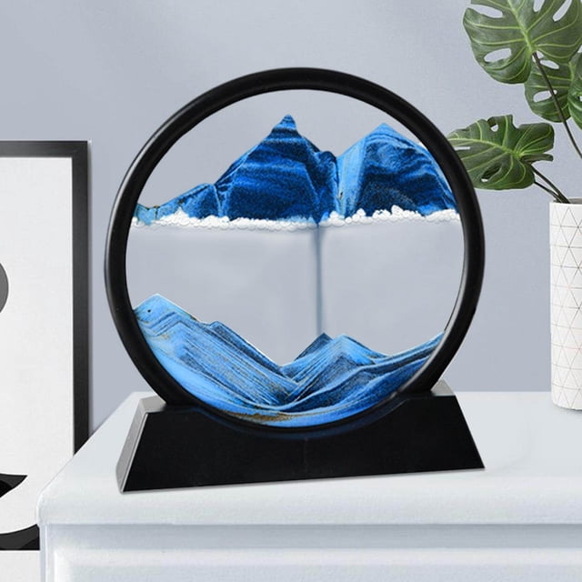 Langgg Tableau Artistique de Sables Fluides Mettant en Vedette un Paysage de Sable Rond et Dynamique en Mouvement avec du Sable Bleu et Blanc Créant un design Superposé Façon Montagne. la Pièce Est Encadrée dans un Cadre Circulaire Noir avec une base Robuste, Affichée sur un Bureau Blanc aux Côté d'Éléments de Déco Minimalistes.