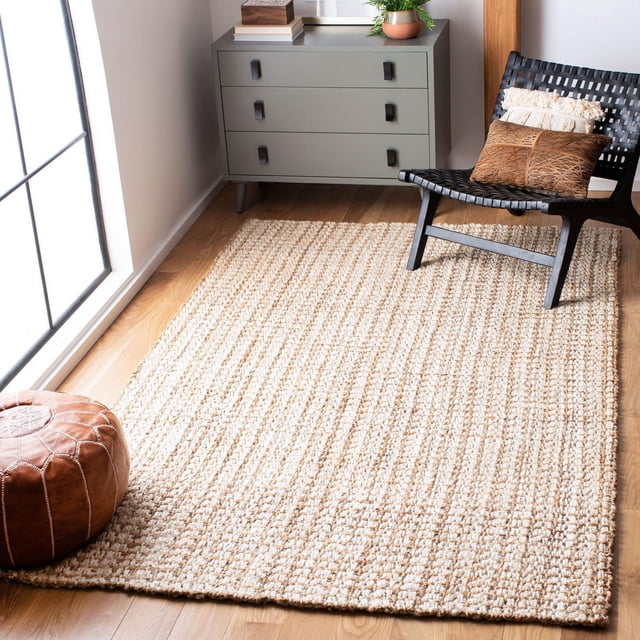 Safavieh Natural Fiber Aletha Solid Area Rug with a woven light beige sisal texture on a hardwood floor, placed near a black woven chair with decorative pillows and a brown leather pouf by a window, accompanied by a gray three-drawer cabinet with plants and books, suitable for coastal or casual interiors.
