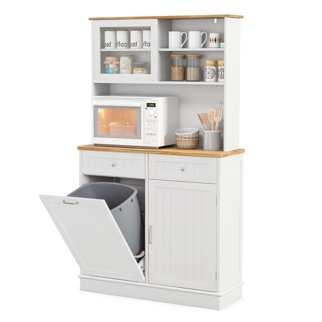Gymax Double Tilt Out Trash Cabinet with Hutch in white featuring a natural rubberwood countertop. This kitchen cabinet includes a tilt-out trash compartment for 10-gallon bins, two drawers, a side cabinet, and an upper hutch with tempered glass doors and open shelving. Designed for pantry organization, it offers cable management holes and adjustable shelves for versatile storage. The cabinet is shown with kitchen essentials like mugs, jars, and a microwave placed on the countertop.