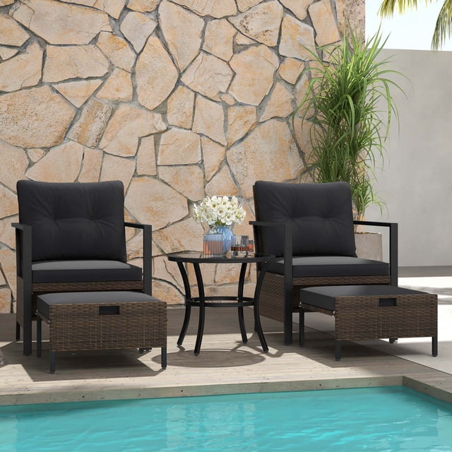 Costway 5 PCS Outdoor Furniture Set featuring two rattan armchairs with black cushions, two multifunctional ottomans, and a round coffee table with a tempered glass top. The set is crafted with mix brown PE rattan and a sturdy metal frame, designed for durability and style. The ottomans can be used as footrests, side tables, or additional seating and are conveniently stored under the chairs. The coffee table is adorned with decorative items and positioned on a wooden deck near a pool, with a stone wall and greenery in the background.