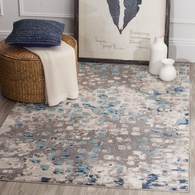 Safavieh Monaco Driskoll Abstract Area Rug with a durable polypropylene power-loomed construction and a mix of high and low loop pile, displaying an erased-weave abstract pattern in blue, grey, and cream tones on a light wood floor, styled with a woven ottoman holding a blue cushion and cream throw, and two white ceramic vases beside a framed botanical print, ideal for adding Bohemian-chic flair to home décor.