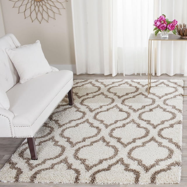 Safavieh Hudson Louise Geometric Shag Area Rug featuring a thick ivory shag pile with brown onion-shaped tile patterns, placed in a living room with a white upholstered sofa and a gold-legged side table with pink flowers, ideal for adding texture to modern or urban décor.