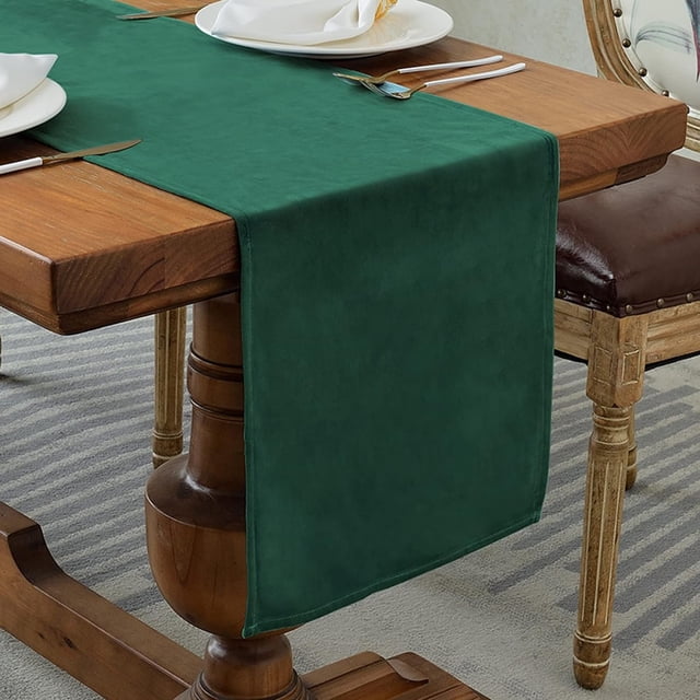 XINQIHANG Velvet Table Runner in dark green, measuring 14x95 inches, displayed on a wooden dining table. The rustic-style table runner is made of 100% velvet, offering a smooth texture and stain-resistant properties. Suitable for dining room, kitchen, or party décor, it complements various table shapes, including rectangular, round, and square. The setting includes a neatly arranged dining setup with plates and cutlery.