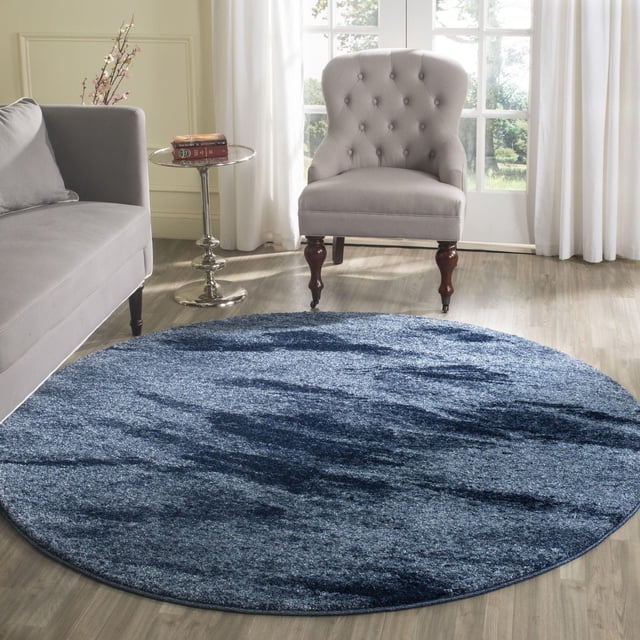Safavieh Retro Panit Abstract Area Rug, a round low-pile polypropylene rug with deep blue and navy abstract patterns, placed on light wood flooring in a bright room with a grey tufted armchair, grey sofa, and a small round metal side table with books and flowers, ideal for mid-century modern accent decor.