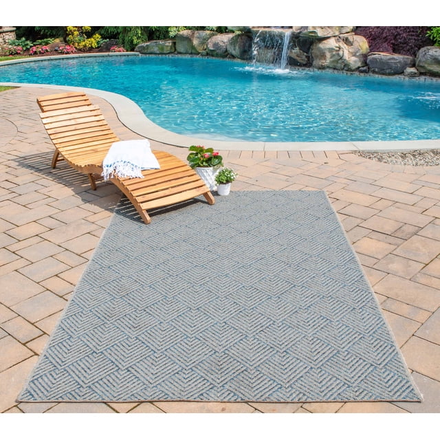 ECARPET Laura Contemporary Area Rug with a repeating geometric pattern in azure blue and off-white tones, made from stain-resistant polypropylene fibers, placed on a stone patio beside a curved outdoor pool with a waterfall, accompanied by a wooden lounge chair and potted plants, suitable for indoor and outdoor use.