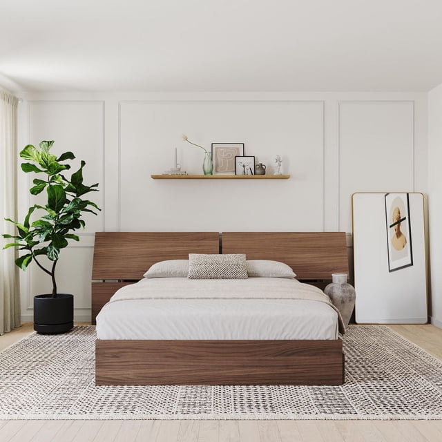 Nexera Queen Size Panoramic Headboard with a low, wide profile in natural walnut finish, displayed in a minimalist bedroom with beige bedding and a patterned lumbar pillow. The headboard pairs with a matching bed frame on a textured area rug, beside a large potted plant and a full-length mirror with abstract art. Suitable for modern bedroom updates and compatible with Nexera queen beds.