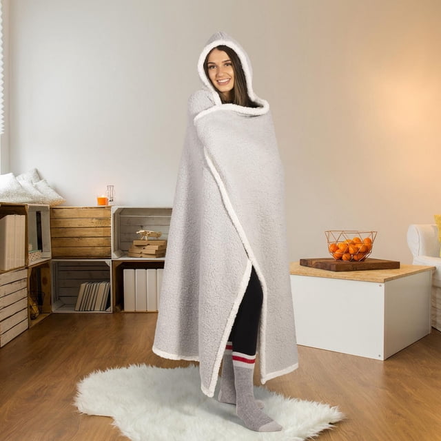Safdie & Co. Premium Ultra Soft Reversible Sherpa Hoodzy Hooded Throw Blanket Light Grey worn by a person standing on a white faux fur rug in a living room. The oversized light grey sherpa blanket with white trim features a large hood and built-in triangular mitts for full-body warmth. The background includes wooden crate shelves with books and decor, a white coffee table with a wire basket of oranges, and warm ambient lighting. This cozy hooded throw is also suitable for relaxing on the couch or as a stylish accent piece in the bedroom.