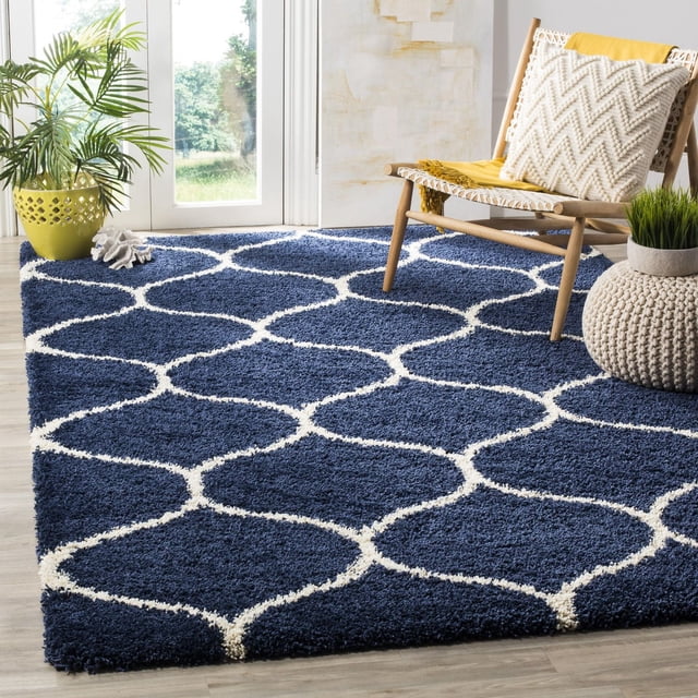 Safavieh Hudson Tapis Shag Géométrique Arline avec une Épaisse Peluche, pile Shag Bleu Marine et Motif de Carreaux en Forme d'Oignon Blanc, Posé sur un Plancher de Bois Clair dans une Pièce Lumineuse avec une Chaise Tissée Tenant un Oreiller Texturé Crème et un Jeté Jaune, un pouf Tricoté avec une Petite Plante Verte, et un Cache-Pot Jaune Près d'Une Porte en Verre. Idéal pour Ajouter de la texture et du Confort aux Espaces de Vie.