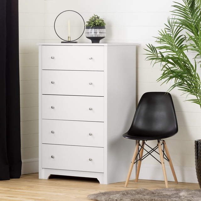 South Shore Vito 5-Drawer Chest for Adult in pure white, a tall upright chest with five spacious drawers featuring metal knob handles and a decorative kick plate. Positioned on a light wood floor against a white shiplap wall, next to a black modern chair with wooden legs and a green potted plant. Its compact design suits bedrooms or craft rooms for organized storage.