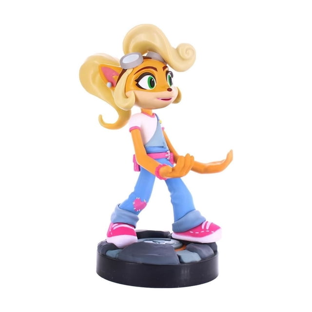 Exquisite Gaming Crash Bandicoot: Coco Cable Guy Phone And Controller Holder is an 8-inch tall figure of Coco Bandicoot wearing blue overalls with a pink patch, white t-shirt with pink trim, and pink sneakers. It stands on a black base with stone-like texture and features large curled blonde hair and outstretched hands to hold phones or gaming controllers compatible with PlayStation, Xbox, and Switch devices.