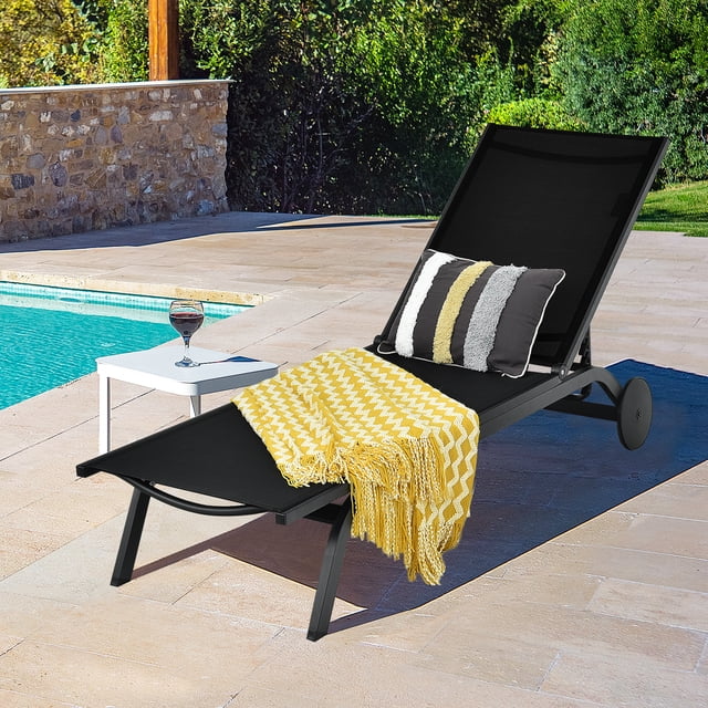 Gymax Chaise Longue de Patio en Aluminium Inclinable Réglable - Ensemble de 1, Noir, Exposée au Bord de la Piscine sur un Patio en Pierre. la Chaise Présente un Cadre en Aluminium Antirouille, un Tissu Noir Respirant et six positions de Dossier Réglables pour un Confort Personnalisé. Deux Roues Arrière Permettent un Mouvement Facile, et une Couverture Jetée à Motif Jaune avec des Coussins Rayés Ajoute une Touche Décorative. une Petite table d'Appoint Blanche avec un Verre de Vin Complète l'Aménagement de Salon Extérieur, Idéale pour Se Détendre au Bord de la Piscine Ou sur un Patio.