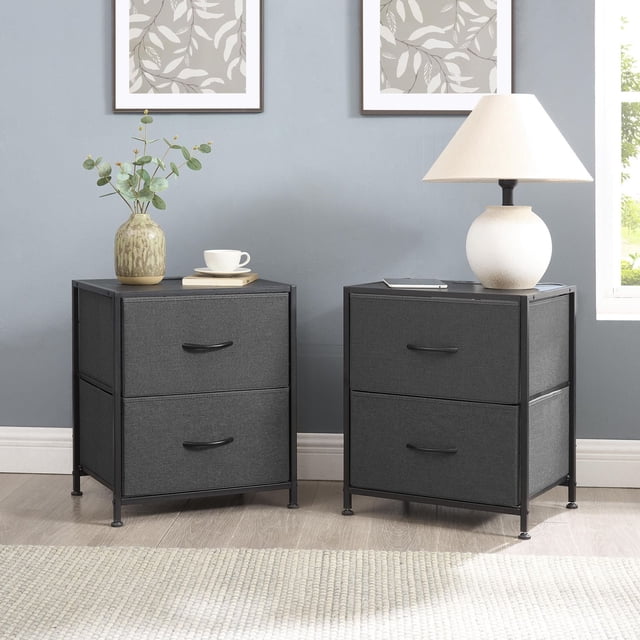 Mainstays 2-Pack Fabric Nightstand set with two grey fabric drawers in a black steel frame, 21 inches tall with adjustable legs, displayed in a bedroom with a lamp, vase, and framed leaf artwork, ideal for compact bedside storage and versatile use.
