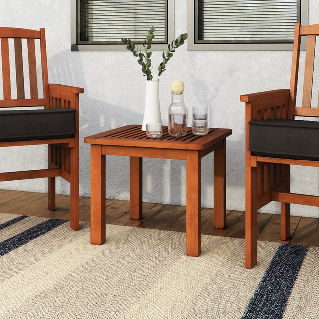 CorLiving Miramar Hardwood Outdoor Square Side Table in brown with a slatted wood top, placed between two hardwood patio chairs with black cushions on a wooden deck. The weather-resistant table holds a white vase with eucalyptus branches, a glass carafe with cork stopper, and two glasses, set against a light grey stucco wall with two windows. Its lightweight, durable design is ideal for small patios and also suitable as an indoor accent table.