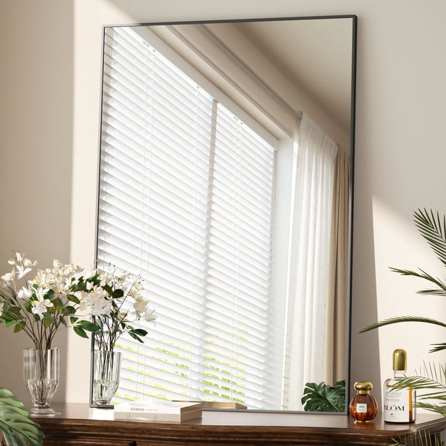 Beautypeak 26"x38" bathroom mirror with rectangular black metal frame, displayed leaning against a wall. The mirror reflects a window with white blinds and sheer curtains, surrounded by decor including vases with white flowers and small bottles on a wooden surface. Sleek and modern wall-mounted design suitable for bathrooms, bedrooms, or living rooms.