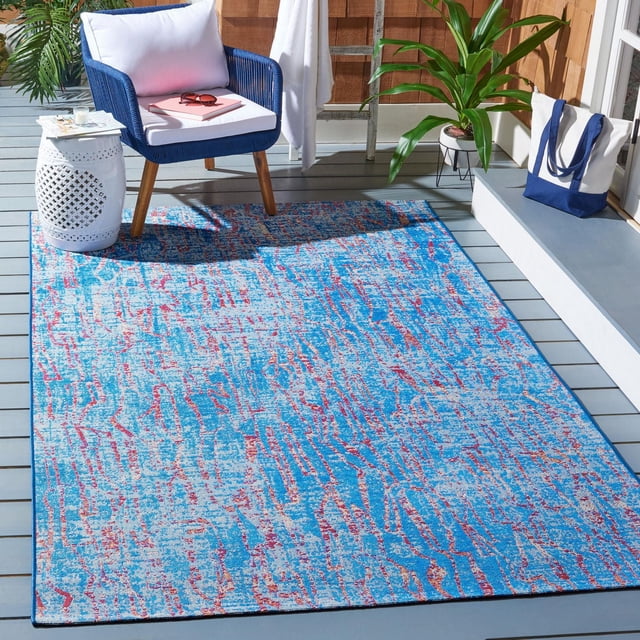 Safavieh Summer Christabella Abstract Outdoor Area Rug with blue, red, and light grey abstract pattern on a grey wooden deck, styled with a blue woven armchair with white cushions, white ceramic side table, green potted plant, and white-blue tote bag, suitable for patios and backyard decks.