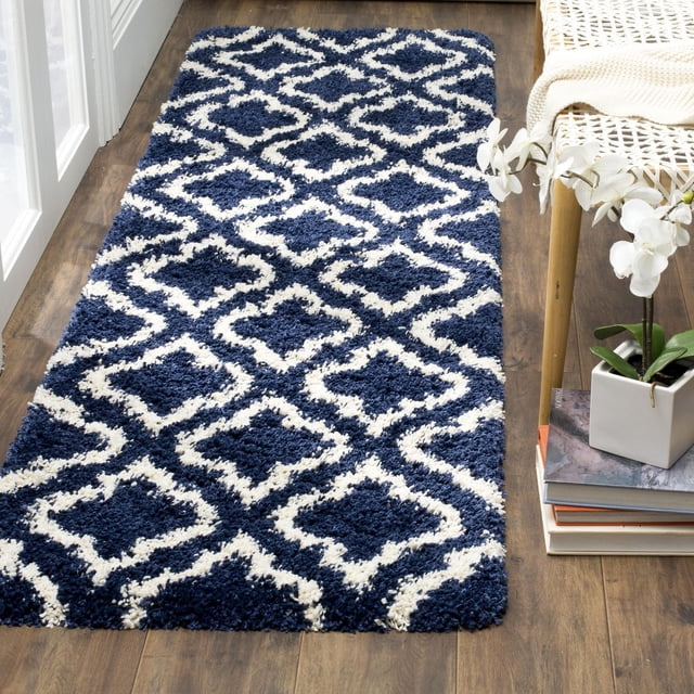 Safavieh Hudson Louise Geometric Shag Area Rug with a thick plush shag pile in navy blue and ivory geometric tile patterns, shown on a hardwood floor next to a woven chair and a white orchid plant, suitable for adding texture to urban or modern décor.
