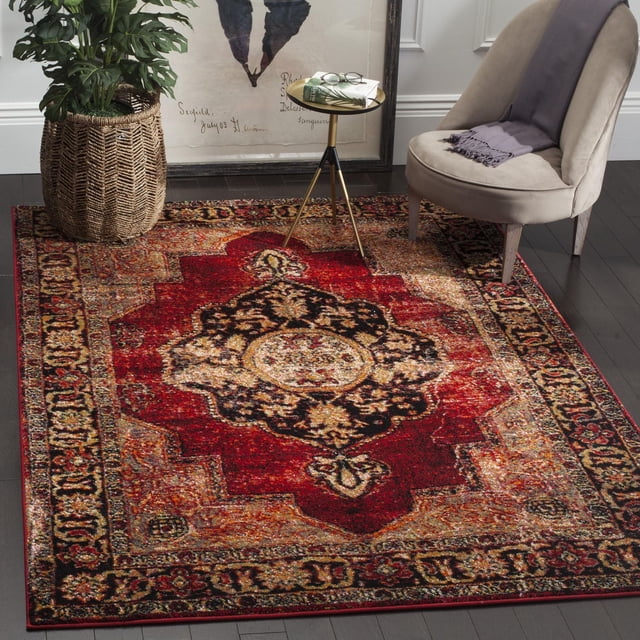 SAFAVIEH Vintage Hamadan Tahnee Traditional Area Rug with a Persian-style medallion pattern in red, black, and beige tones and antique patina finish, placed on dark hardwood flooring. Styled with a beige accent chair draped with a gray throw, a small gold tripod side table holding books and glasses, and a woven basket planter with a green leafy plant, ideal for adding vintage charm to living rooms or reading nooks.