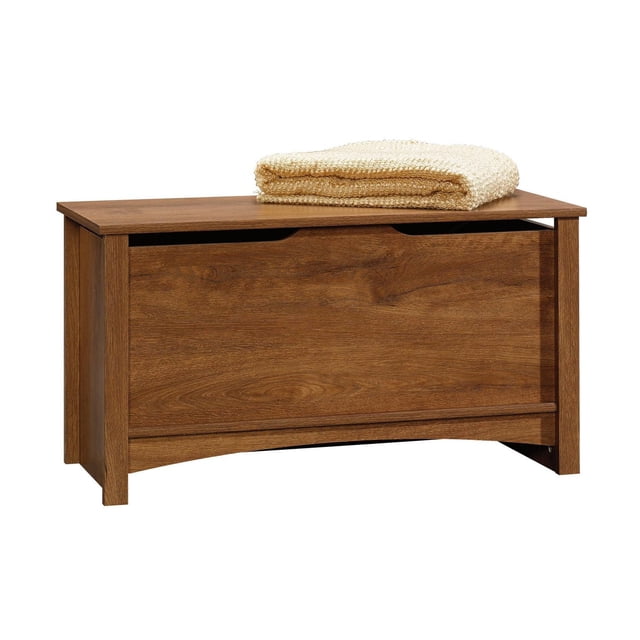 Sauder® Shoal Creek Collection Storage Chest in a warm wood finish, featuring a rectangular design with sculpted edges, tapered legs, and Spanish-finish hardware. The chest is shown with a folded beige blanket on top, highlighting its versatile use for storing blankets, pillows, and other items. Equipped with standard ventilation and a lid stay for safety, this storage chest is suitable for living rooms, bedrooms, or offices.