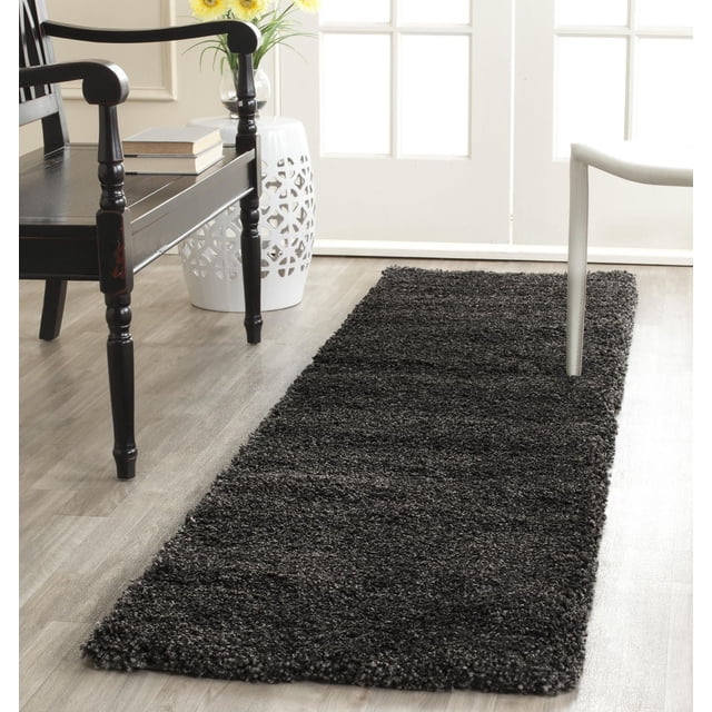 Safavieh Milan Harlow Solid Shag Area Rug in dark charcoal gray with dense shag texture, rectangular on light wood flooring between a black wooden armchair and a white modern chair, beside a white ceramic garden stool with cutout patterns and a glass vase with yellow flowers by a bright window, suitable for adding warmth and texture to contemporary home décor.