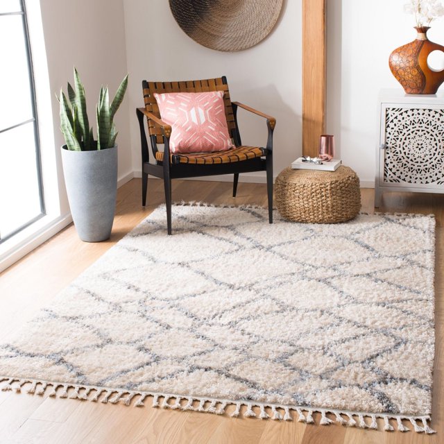 SAFAVIEH Berber Fringe Felicia Geometric Shag Area Rug in a bright room on light hardwood floors, featuring a beige shag rug with gray diamond geometric patterns and fringed edges. The rug is styled with a woven leather strap chair with a pink geometric cushion, a round woven ottoman with a book and glass, a tall gray planter with snake plants, and a decorative woven wall hanging.