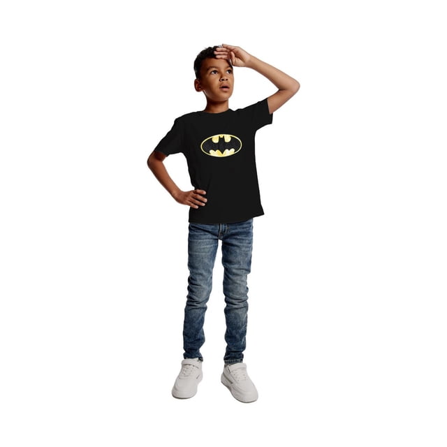Boys Batman Logo T-Shirt in black with a yellow Batman emblem in HD Gel and Puff finish on the front, paired with blue denim jeans and white sneakers, shown in a full-body front view with one hand on hip and the other raised to forehead.