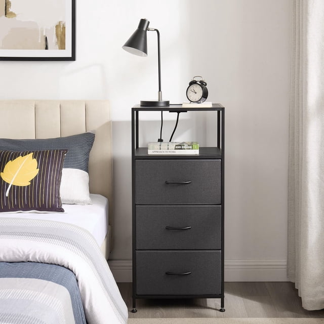 Mainstays 3 Drawer Fabric Nightstand with USB in grey fabric and black metal frame, featuring three removable fabric drawers with black handles and an open shelf holding books, topped with a black desk lamp and alarm clock, placed beside a beige upholstered bed in a bedroom with light walls and curtains, ideal for compact bedroom storage and organization.