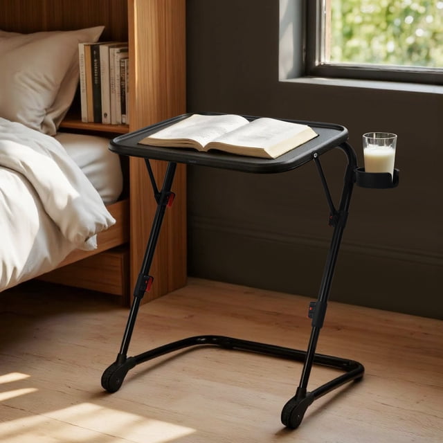 Homy Casa Foldable Laptop Table with Fixed Cup Holder and Adjustable Metal Frame featuring a matte black plastic tabletop holding an open book, supported by a foldable powder-coated steel frame with wheels and red locking clips. The fixed cup holder holds a glass of milk. Positioned on a light wood floor near a bed and window, it is ideal for small spaces and also suitable as a snack or reading table.