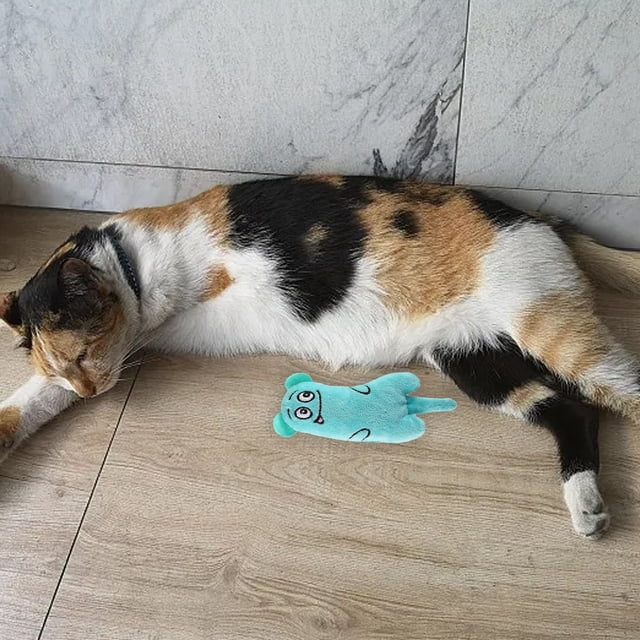 WIFKLSIIPG Since Hi Cat Toy Cute Sound Paper Pet Toy Tail Toy, made of durable polyester with a crinkly sound effect to captivate cats. The toy, in a light blue color with a cheerful face design, is placed beside a calico cat lying on a wooden floor. Suitable for encouraging active play and reducing boredom in cats.