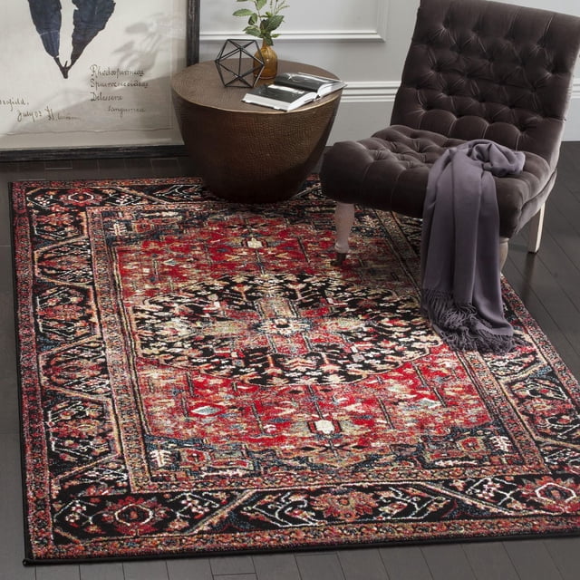 Safavieh Vintage Hamadan Farran Traditional Area Rug with a richly colored Persian-style pattern in red, black, and beige, set on dark hardwood floors beside a tufted dark gray chair with a purple throw and a round bronze side table holding decorative items and an open book, suitable for enhancing vintage charm in modern living spaces.