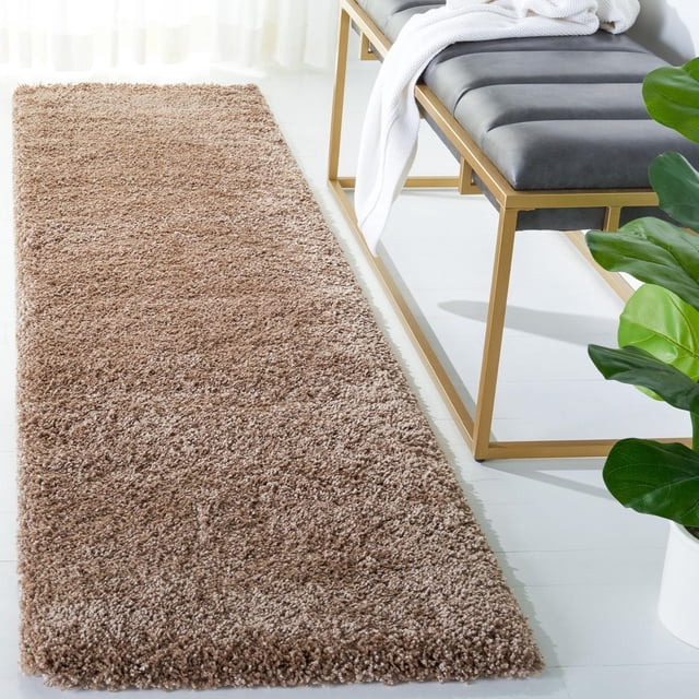Safavieh Milan Harlow Tapis Shag Uni Brun avec une pile de Fil Synthétique dense et Douce, Posé sur un Plancher Blanc à Côté d'Un Banc à Cadre Métallique Doré avec des Coussins Noirs et un Jeté Blanc, à Côté d'Une Plante en Pot Verte, Idéal pour Ajouter Chaleur et texture à la Déco Contemporaine de la Maison.