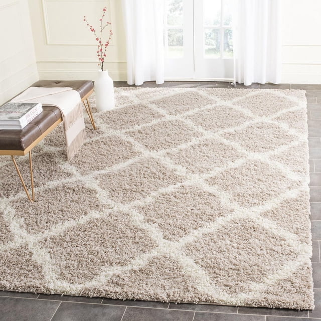 Safavieh Dallas Logan Geometric Shag Area Rug with beige and ivory Moroccan tile pattern on a plush polypropylene shag pile, displayed on gray tile flooring in a bright room with sheer white curtains, a brown leather bench, and a white vase with red berry branches, ideal for adding texture and comfort to home interiors.