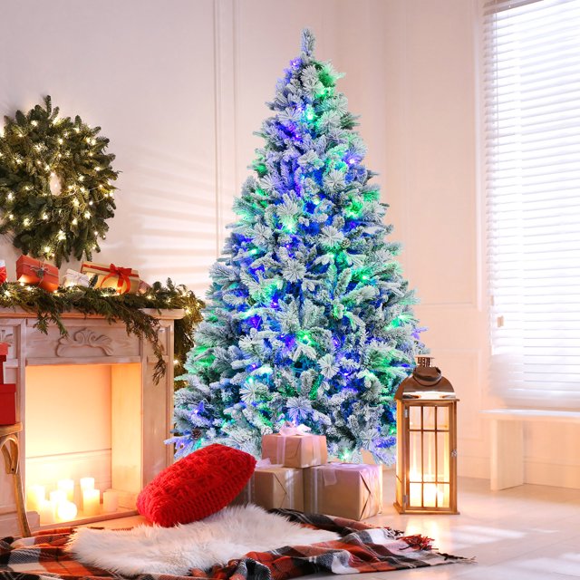 Gymax 7 FT Pre-Lit Artificial Christmas Tree with snow-flocked branches, 320 colorful LED lights, and 1270 branch tips. The tree features a mix of PVC, PE leaves, and pine needles, adorned with 40 pine cones for a natural and festive look. It is displayed in a cozy indoor setting with wrapped gifts, a lit fireplace, and holiday decor, creating a warm and inviting atmosphere.