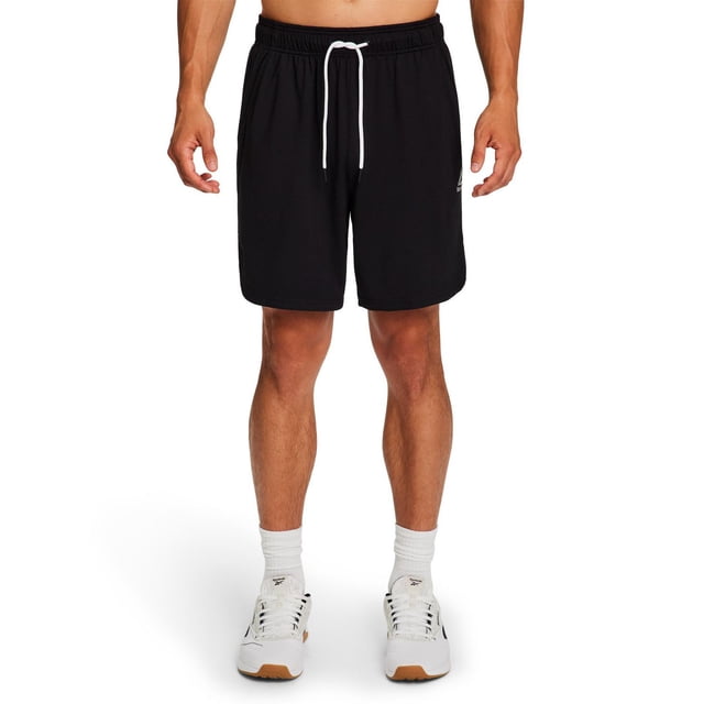 Reebok Men's Delta Core Shorts in black soft jersey with elastic waistband and white drawcord, featuring side pockets and the Reebok Delta logo on the left thigh, worn front-facing with white Reebok sneakers and socks, ideal for workouts or casual wear.