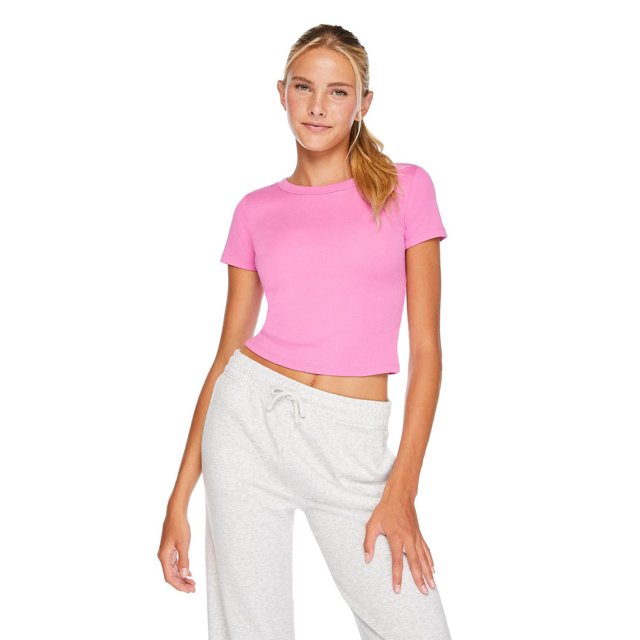 No Boundaries Women's Rib Baby Tee in pink shown front-facing on a model wearing light grey drawstring sweatpants. The rib knit tee features a crew neckline, short sleeves, and a curved hem, offering a fitted silhouette ideal for casual and versatile styling.