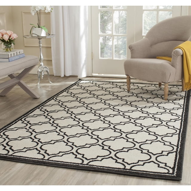 Safavieh Amherst Wilson Tapis Géométrique Mettant en Vedette un Motif de Treillis Noir et Blanc avec une Bordure Sombre, construction en Polypropylène Tissé Mécaniquement pour la Durabilité dans les Contextes Intérieur-Extérieur, Placé sur un Plancher de Bois Clair à Côté d'Un Fauteuil beige avec un Jeté Jaune, Convient aux Zones à Fort Achalandage à la Maison Ou au Bureau.