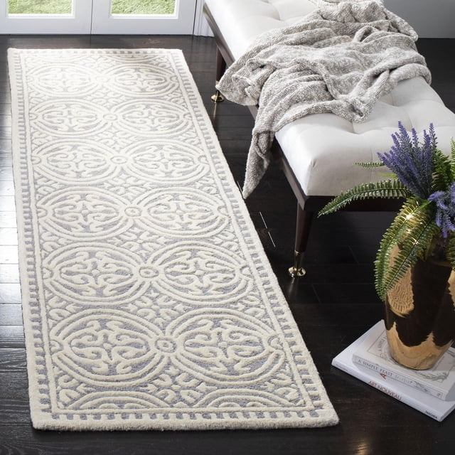 SAFAVIEH Cambridge Leslie Geometric Area Rug with hand-tufted wool featuring a raised geometric circular pattern in light neutral shades, placed on dark hardwood floor beside a white tufted bench with a gray knitted throw and a gold planter with green and purple plants on stacked books, suitable for enhancing hallways or entryways.