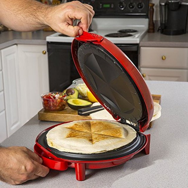 Elite Gourmet Mexican Taco Tuesday Quesadilla Maker in red, an 11-inch small appliance designed for easy-slice 6-wedge quesadillas and grilled cheese. The device is shown on a kitchen countertop, grilling a quesadilla with golden-brown wedges. A hand lifts the lid, revealing the non-stick interior surface. Ingredients like avocado and salsa are visible in the background.