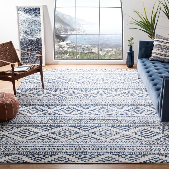 SAFAVIEH Tulum Lakisha Distressed Southwestern Area Rug in a bright living room with a large window showing a rocky coastal view. The rug has a cream background with blue southwestern geometric patterns in a distressed design. It rests on light hardwood floors, near a blue tufted sofa with patterned pillows, a brown woven leather chair, a leather pouf, a tall green plant in a blue vase, and abstract wall art. Made of stain-resistant synthetic fibers with a 0.3-inch non-shedding pile, it suits living rooms, dining rooms, and other home areas.