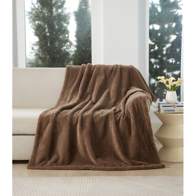 Better Homes & Gardens Faux Fur Throw in taupe, 50 x 72 inches, draped over a beige sofa in a bright room with large windows. This oversized reversible throw features plush faux fur on both sides, ideal for warmth and comfort in living rooms or bedrooms.