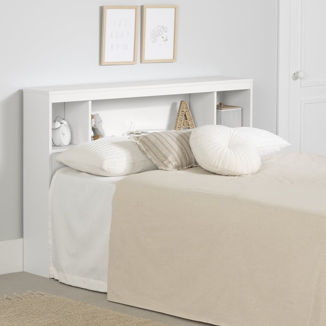 South Shore Spark Full Bookcase Headboard with Storage in pure white, featuring three open storage compartments with fixed shelves and a cable management hole in the back panel. The headboard is styled with a white alarm clock, a small stuffed animal, a woven letter A, and a white basket. It is paired with a bed dressed in beige and white pillows and a beige bedspread against a light grey wall with two framed prints above.