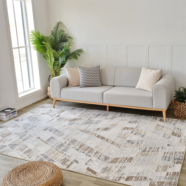 Modern beige and brown abstract indoor area rug placed beneath a light gray sofa with chevron and textured pillows, complemented by green plants in woven baskets near large windows.
