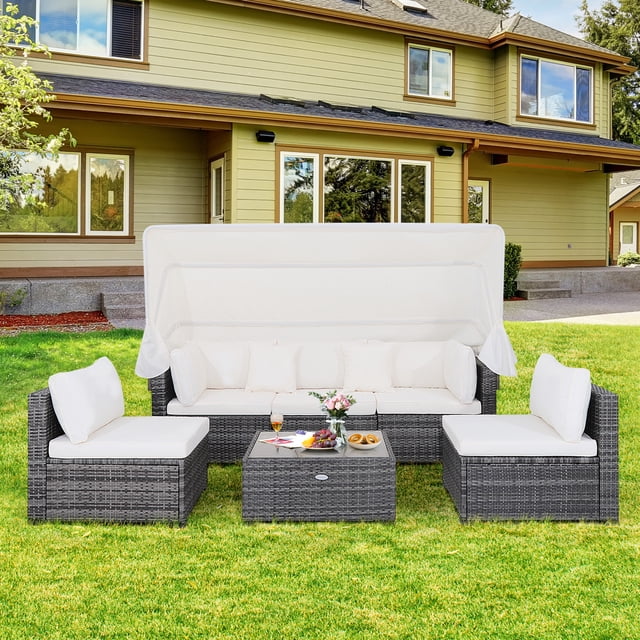 Gymax Ensemble de Mobilier de Patio en Rotin PE 6 Pcs Présentant un design en Rotin Gris Mixte avec des Coussins Blanc Cassé, une table Basse en Verre Trempé et un Auvent Rétractable. l'Ensemble Comprend un sofa 3 Places, Deux sofas d'Angle, Trois sofas Sans Accoudoirs et Deux Coussins Décoratifs, Disposé sur une Pelouse Verte devant une Maison. l'Auvent Est Allongé, Offrant de l'Ombre pour l'Aire d'Assise.