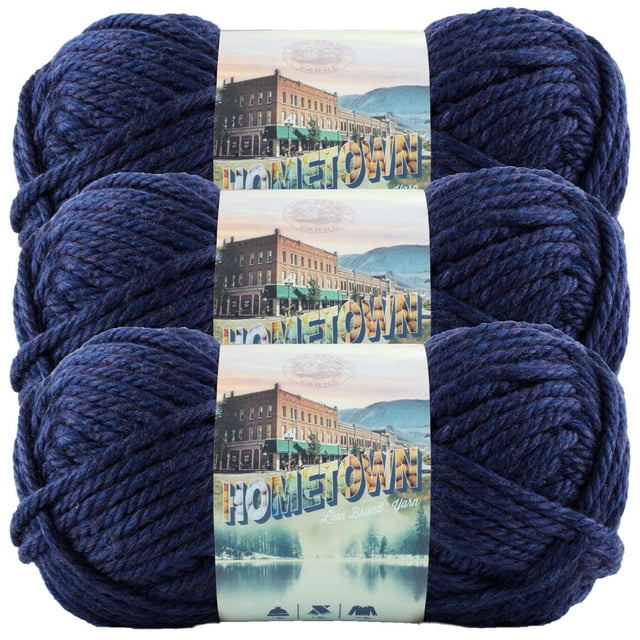 Lion Brand Hometown Yarn - Blue Sky featuring three skeins of thick, textured deep blue yarn stacked vertically with labels depicting a small town street and mountain backdrop. This machine washable yarn is suitable for fast knit and crochet projects.